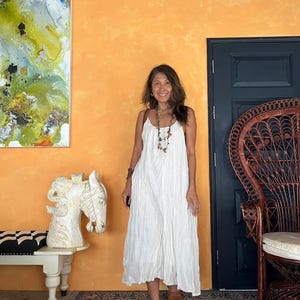 May include: A woman wearing a white, flowing sundress with thin straps and brown sandals. She is standing in front of an orange wall with a dark blue door and a decorative chair. A painting and a white horse sculpture are also visible.