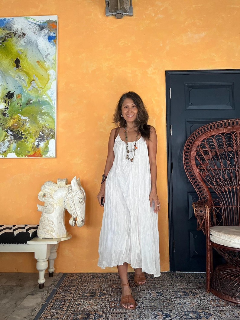 May include: A woman wearing a white, flowing sundress with thin straps and brown sandals. She is standing in front of an orange wall with a dark blue door and a decorative chair. A painting and a white horse sculpture are also visible.