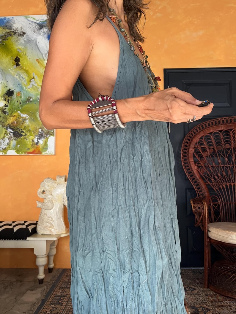 May include: A light blue dress with a textured, pleated design. A wide, silver-toned bracelet with red, white, and gray accents is worn on the left wrist. The background includes an orange wall, a painting, and a wicker chair.