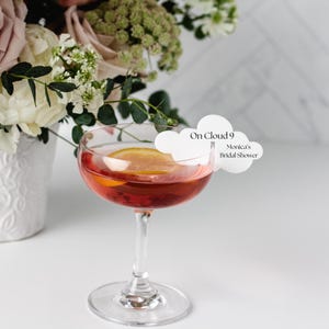 May include: A white cloud-shaped card with the text "On Cloud 9" and "Monica's Bridal Shower" on a red cocktail in a coupe glass. The glass is sitting on a white surface with a bouquet of pink and white flowers in a white vase in the background.
