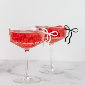 May include: Two clear glass champagne flutes filled with a red liquid and garnished with white and black ribbon bows.