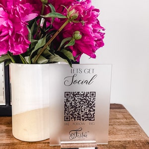 May include: A frosted acrylic sign with a black QR code and the text "Let's Get Social" and "Like Follow Tag". The sign also has the text "Festive" in a cursive font. The sign is sitting on a wooden table with a bouquet of pink peonies in a white vase.