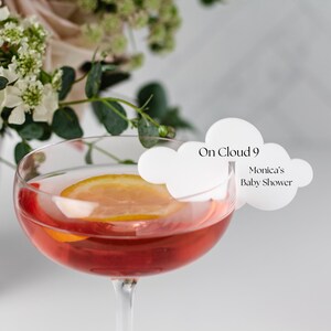May include: A white cloud-shaped drink stirrer with the text "On Cloud 9" and "Monica's Baby Shower" printed on it. The stirrer is placed in a glass of red liquid with a lemon slice.