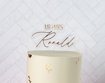 May include: A wedding cake with gold leaf accents and a clear topper that reads "MR + MRS" and the name "Ronald" in gold script. The cake is on a white surface with a pink cloth.