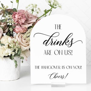 May include: White sign with black text that reads "The drinks are on us! The hangover is on you! Cheers!" The sign is shaped like an arch and is sitting on a white surface.