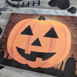 May include: A paper craft pumpkin with a black smiling face and triangle eyes. The pumpkin is orange and is set against a dark brown background with black trees and a graveyard.