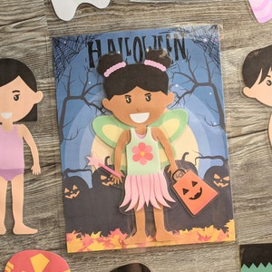 May include: A paper doll craft set featuring a fairy with brown skin, pink wings, and a green dress with a flower. The doll is holding a wand and a pumpkin-shaped bag. The background is a blue night sky with a moon and trees. The text "HALLOWEEN" is at the top of the image.