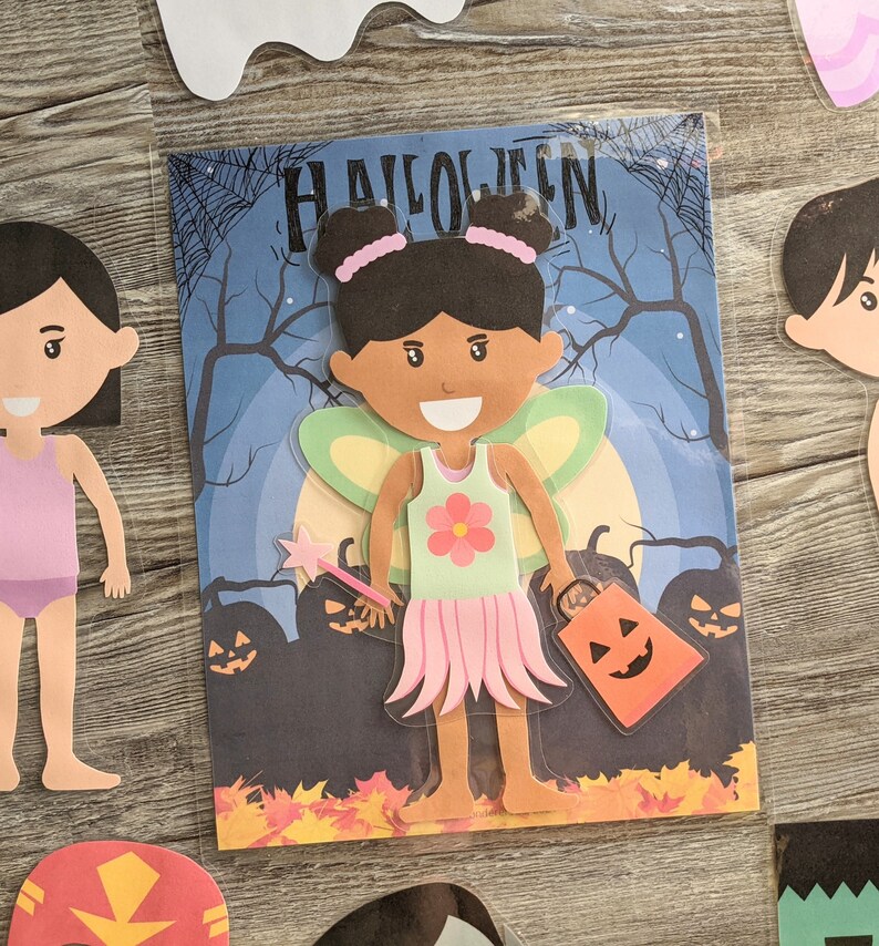 May include: A paper doll craft set featuring a fairy with brown skin, pink wings, and a green dress with a flower. The doll is holding a wand and a pumpkin-shaped bag. The background is a blue night sky with a moon and trees. The text "HALLOWEEN" is at the top of the image.