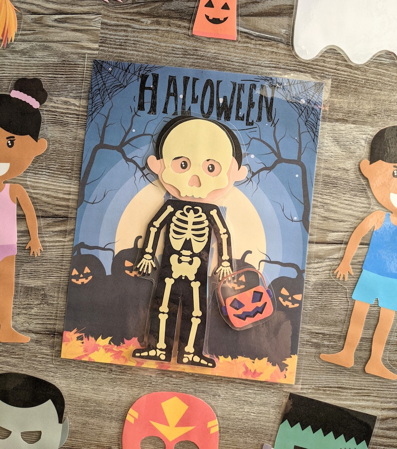 May include: A Halloween-themed paper craft activity featuring a skeleton character holding a pumpkin-shaped bucket. The character has a yellow skull face and is dressed in black pants and a skeleton shirt. The background is a blue night scene with a full moon and pumpkins.