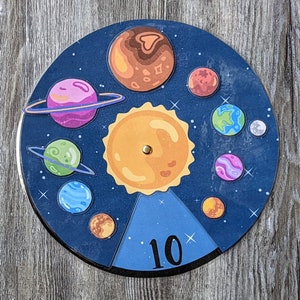 May include: A blue and black circular game board with a yellow sun in the center and 10 planets surrounding it. The planets are different colors and sizes. The board has the number 10 printed on it.
