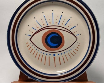 May include: A white ceramic plate with a brown and blue rim. The center of the plate features a hand-painted design of a blue eye with a crescent moon inside. The eye is surrounded by a radiating pattern of brown and blue lines.