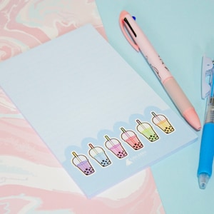 Pode incluir: Um bloco de notas azul claro com ilustrações coloridas de bubble tea. Uma caneta multi-cor rosa e uma caneta azul estão colocadas ao lado do bloco de notas em uma superfície azul claro e rosa. O bloco de notas tem o texto "AVA KIRA".