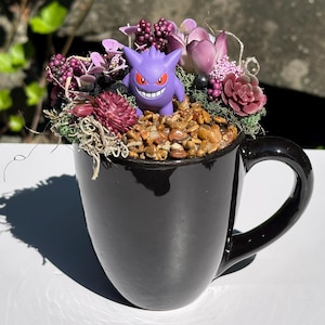 May include: A black ceramic mug filled with a decorative arrangement. The arrangement includes a purple Pokemon figure, dried flowers, and small stones. The flowers are various shades of purple and burgundy. The mug has a large handle and sits on a white surface.