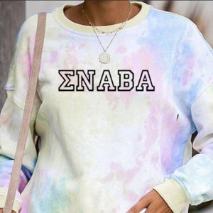 Könnte beinhalten: Ein tie-dye Sweatshirt mit langen Ärmeln und einem Rundhalsausschnitt. Das Sweatshirt ist hellblau, rosa und gelb auf weißem Hintergrund. Der Text "ΣNABA" ist in Schwarz auf der Vorderseite des Sweatshirts aufgedruckt.
