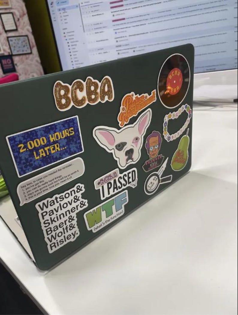 May include: A green laptop covered in stickers. The stickers include a dog, a pear, a magnifying glass, a record, a  '2,000 hours later' sticker, a 'BCBA I Passed' sticker, and a 'WTF (what's the function)' sticker.