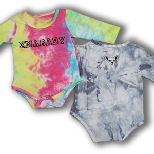 May include: Two long-sleeved baby onesies. One is tie-dye in pink, yellow, and blue with the text "ΣNABABY" in black. The other is tie-dye in gray and white with a black and white illustration of a dog's head.