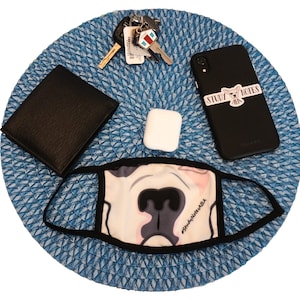 May include: A blue and white patterned face mask with the text "#StudyNotesBabe" printed on it. A black leather wallet, a white AirPod case, a black phone with a sticker that says "Study Notes", and a set of keys are on a blue and white woven mat.