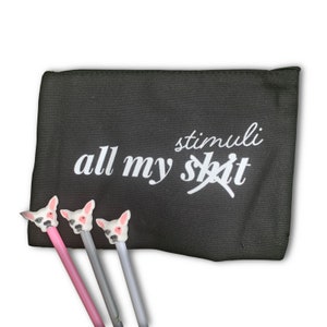 May include: Black canvas zippered pouch with the text "all my stimuli" in white. Three pens with dog head toppers in pink, gray, and silver are in front of the pouch. The word "shit" is crossed out.