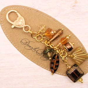 May include: A gold-colored charm with a heart-shaped clasp and a collection of amber, brown, and green beads. The beads are various shapes, including faceted squares, teardrops, and oblongs. The charm is attached to a brown, oval-shaped tag.