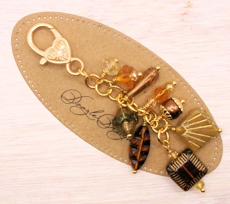 May include: A gold-colored charm with a heart-shaped clasp and a collection of amber, brown, and green beads. The beads are various shapes, including faceted squares, teardrops, and oblongs. The charm is attached to a brown, oval-shaped tag.