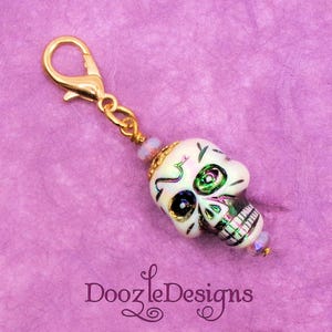 May include: A gold charm with a white skull bead with green and pink details. The skull bead has a painted design of a skull with a single eye and a mouth. The charm is attached to a gold chain with a lobster clasp.