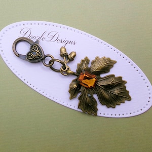 May include: A bronze-toned metal keychain charm featuring a heart-shaped lobster clasp, two small acorn charms, and a large maple leaf pendant accented with a rectangular amber-colored crystal. The charm is displayed on a white oval card with the text Doozle Designs.