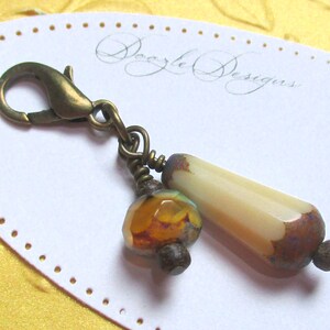 May include: A decorative charm with a lobster clasp. The charm features a faceted orange and green bead, a cream-colored elongated bead with brown accents, and antique brass-colored metal components. The charm is attached to a white card with the text "Dazzle Designs".
