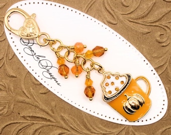 May include: A gold-toned metal keychain featuring a heart-shaped lobster clasp and a charm shaped like an orange mug with a pumpkin design and white foam topped with small orange crystals, accented by dangling orange faceted glass beads.