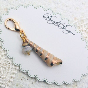 May include: Gold charm featuring a seashell with a copper-colored finish and a small white bead. The charm is attached to a gold lobster clasp.