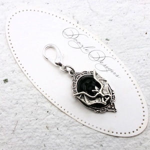 May include: Silver charm with a black enamel bat design in an ornate frame. The charm has a lobster claw clasp.