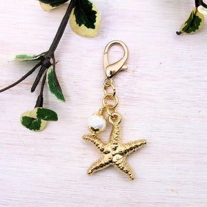 May include: Gold starfish charm with a white pearl bead and a gold lobster clasp. The charm is hanging from a gold chain.