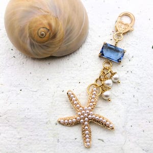 May include: A gold-colored starfish charm with pearl accents and a blue rectangular gem. The charm is attached to a gold clasp and is next to a seashell. The charm is a beach-themed accessory.