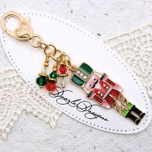 May include: A gold-colored keychain with a heart-shaped clasp and a decorative charm. The charm features a colorful, detailed nutcracker figure with red, green, and pink accents, and dangling beads. The text "Doogle Designs" is visible.