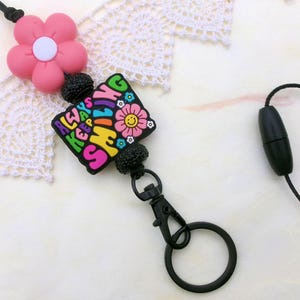 May include: A pink flower and a black lanyard with a charm that says "ALWAYS KEEP SMILING" in colorful letters. The charm has a flower with a smiling face. The lanyard has a black clasp and a black ring.