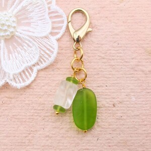 May include: A gold-colored clasp with a chain holding two green and clear glass beads. The beads are oval and rectangular. A white lace flower is in the background.