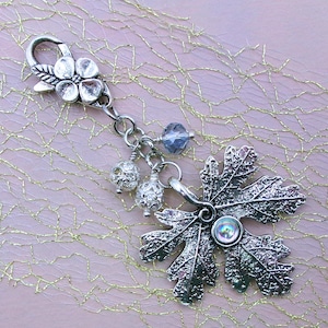 May include: A silver-toned metal charm featuring a textured oak leaf pendant with a central iridescent cabochon. The charm includes a lobster clasp adorned with a small flower, two spherical filigree beads, and a single faceted blue crystal bead.