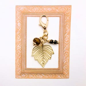 May include: A gold-colored leaf charm with a lobster clasp, adorned with brown beads and a small gold bead. The charm is displayed against a peach-colored decorative frame with a white background. The leaf charm is a jewelry accessory.