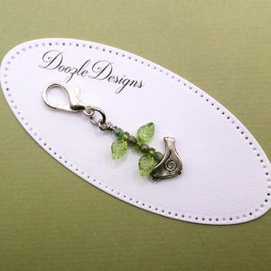 May include: A silver-toned metal lobster clasp charm featuring a small bird figure with a spiral design, accented by two translucent green leaf-shaped beads and small green round beads, attached to a white oval display card labeled Doozle Designs.