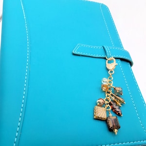May include: A turquoise blue planner with a matching strap closure and white stitching. A gold-colored charm with various beads and decorative elements hangs from the strap. The charm includes brown, gold, and clear beads.