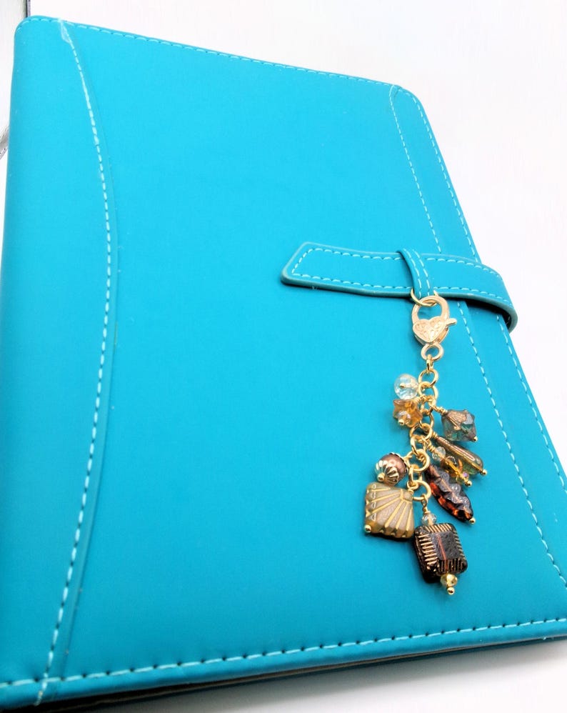 May include: A turquoise blue planner with a matching strap closure and white stitching. A gold-colored charm with various beads and decorative elements hangs from the strap. The charm includes brown, gold, and clear beads.