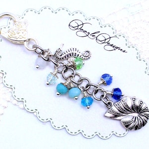 May include: A silver charm bracelet with a seahorse, seashells, and turquoise and blue beads. The charm bracelet is attached to a silver lobster clasp. The bracelet is on a white background with the text "Doodle Designs".