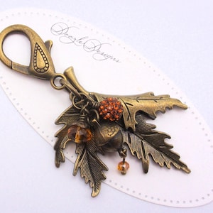 May include: A bronze-colored key chain with a charm featuring a cluster of oak leaves, acorns, and orange-colored rhinestones. The charm is attached to a lobster clasp.