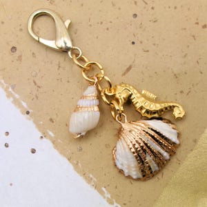 May include: A gold-colored keychain with three charms: a white and gold seashell, a gold seahorse, and a white and gold scallop shell. The keychain has a lobster clasp for easy attachment.