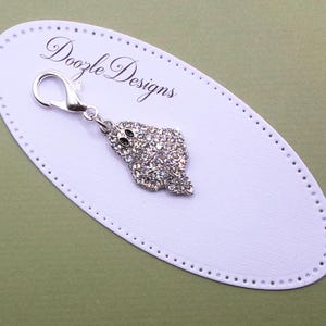 May include: A silver-toned metal charm in the shape of a ghost, covered in clear sparkling rhinestones with two black oval eyes. The charm features a lobster claw clasp and is attached to an oval white display card printed with Doozle Designs.