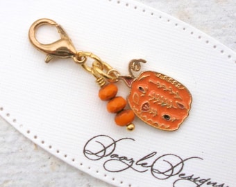 May include: A gold-colored charm with a pumpkin design. The orange pumpkin charm has a gold outline and is connected to a lobster clasp. Three orange beads and a gold bead hang below the pumpkin. The charm is on a white ribbon with the text "Doozle Designs."