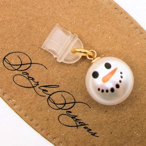 May include: A white pearl snowman charm with black eyes, a black mouth, and an orange nose. The charm is attached to a gold loop and a clear plastic clip.