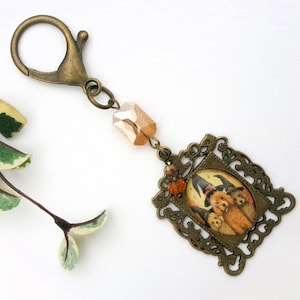 May include: A bronze-toned Halloween-themed keychain. It features a decorative frame with a picture of three dogs wearing witch hats. The keychain has a lobster clasp, a faceted bead, and small orange beads.