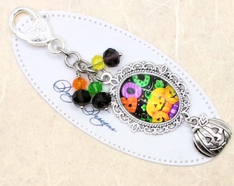 May include: A Halloween-themed keychain with a silver-tone clasp and decorative beads in orange, green, yellow, and purple. The main charm features a silver filigree frame with a colorful pumpkin design. A small pumpkin charm dangles below.