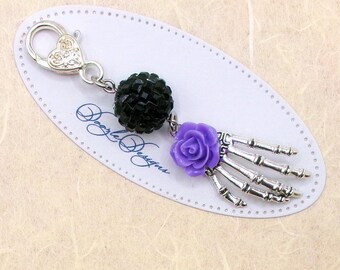 May include: A silver charm with a black beaded skull, a purple flower, and a silver skeleton hand charm. The charm is attached to a silver lobster clasp.