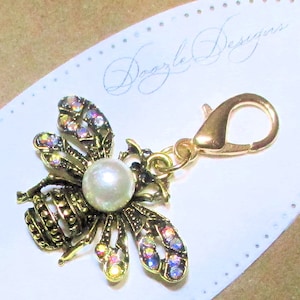 May include: A gold-tone bee charm with iridescent rhinestones and a white pearl. The charm is attached to a gold-tone lobster clasp.